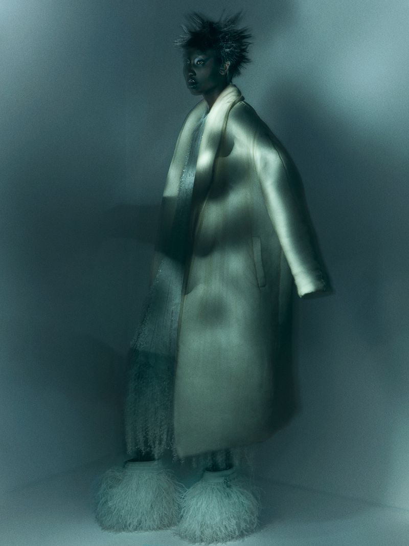 Fashionable woman with spiky hair wearing a beige coat and furry boots posing indoors against a muted blue-gray background.