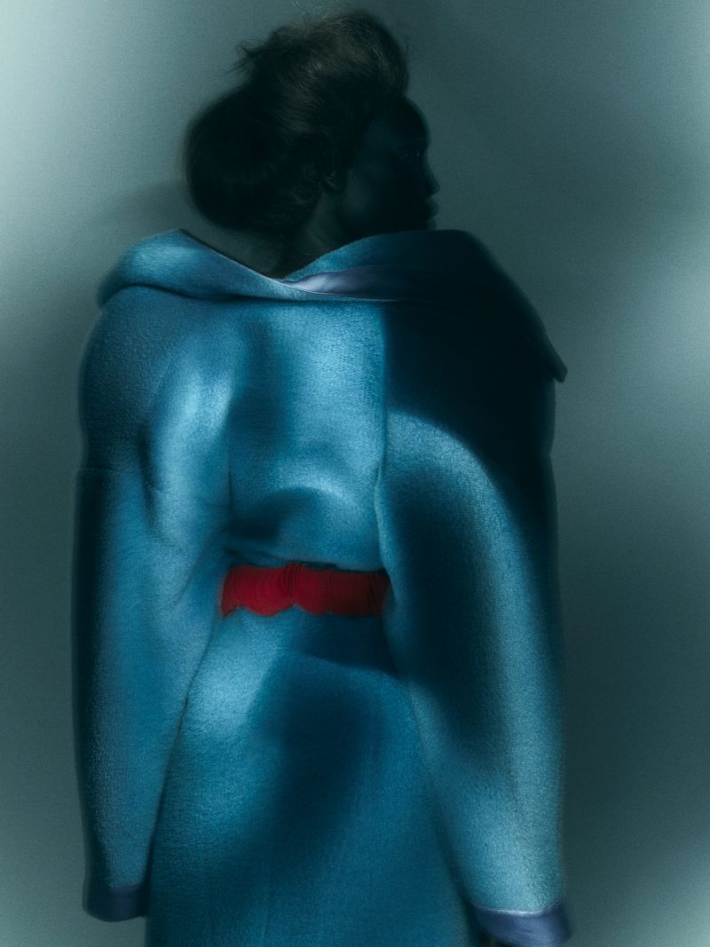 From the back, an adult female with dark hair is wearing a light blue coat with a red sash against a solid, gradient teal background.