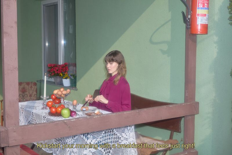 Woman in magenta blouse sits at a lace-covered table with breakfast, including eggs, tomatoes, and apples, on an outdoor porch.