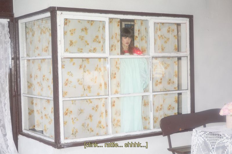 Indoor portrait of a woman standing behind a window with patterned curtains in a red top and light blue dress.