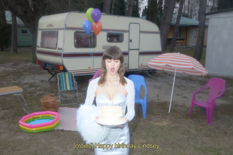 Woman in white dress holding cake outdoors with balloons and camper in background, in vintage style party scene. 