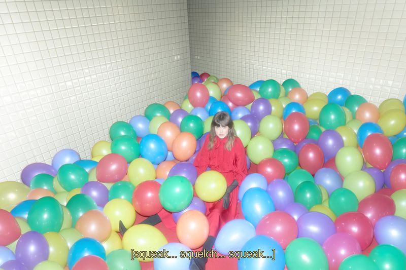 Child in a red jumpsuit sits in a ball pit filled with colorful balloons against a light yellow tiled wall indoors.
