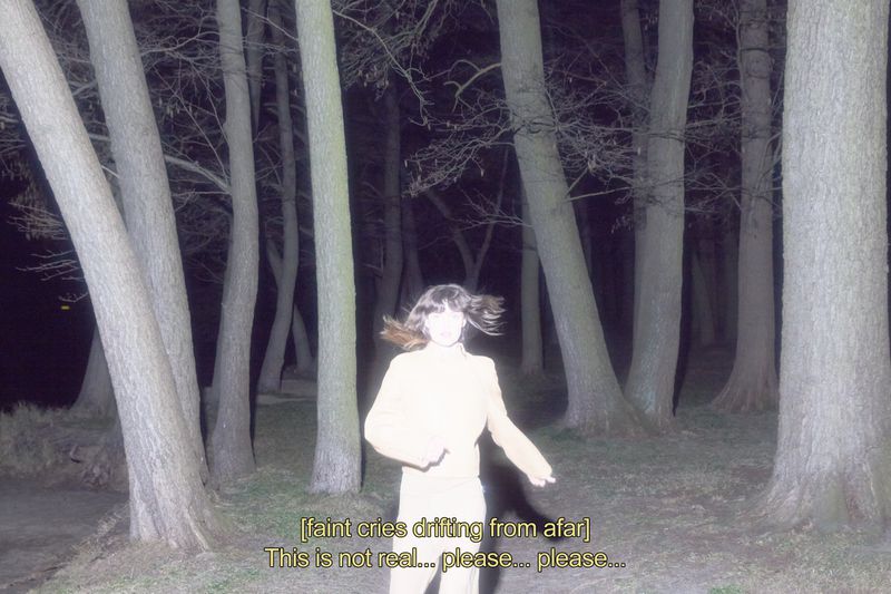 Woman in a light jacket in a dark forest at night. Tall trees surround the individual. Text overlay is visible in the image.