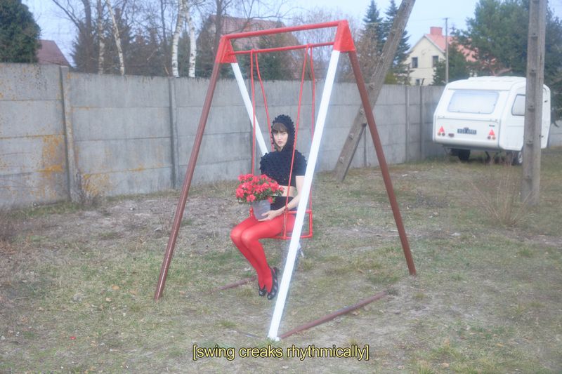 A person in red leggings sits on a red and white swing holding a pot of red flowers outdoors near a cement fence and a white trailer.