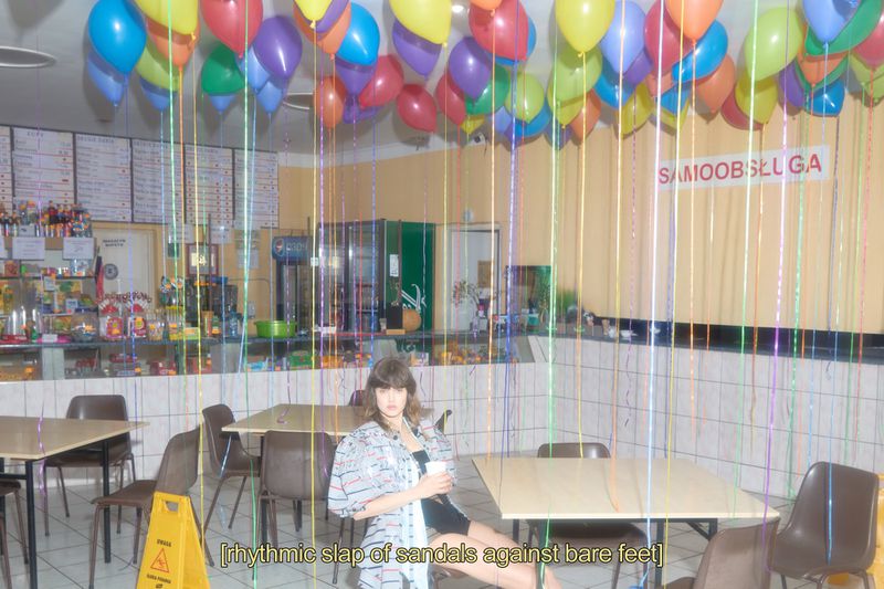 Woman sits at a table in a cafe with balloons overhead, wearing a patterned jacket. Colorful ribbons and balloons decorate the scene.