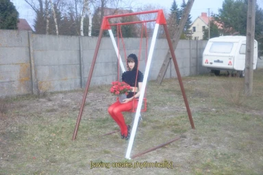 Lindsey Wixson seated on a red playground swing holding a potted plant in an outdoor lot with a caravan and concrete wall