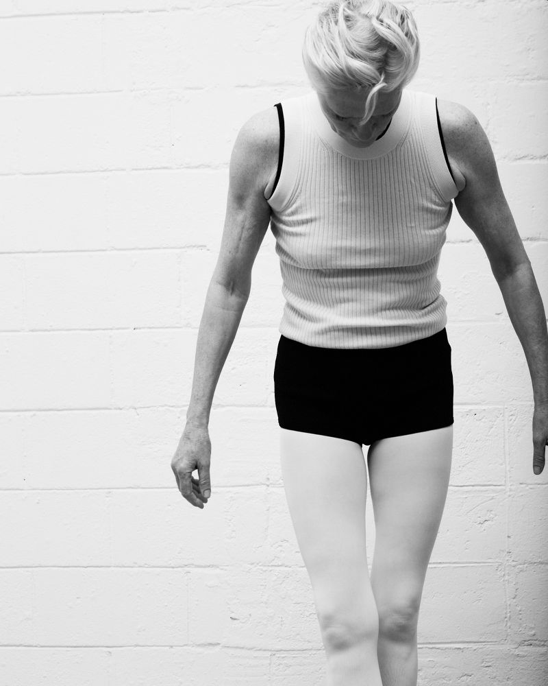 Black and white photograph of an adult female dancer with short blonde hair in tank top and ballet shorts, looking down in a studio.