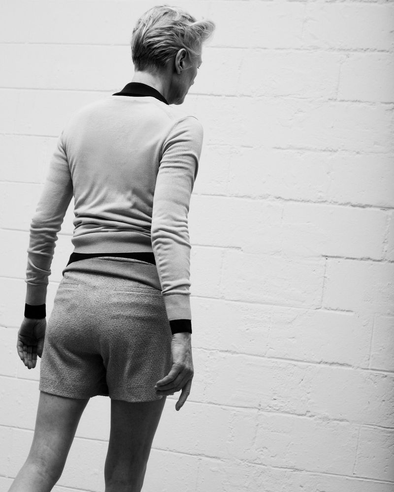 Black and white shot from behind of an adult wearing a light sweater and shorts, standing near a textured white brick wall.