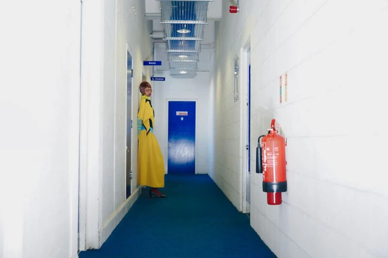 Stylish woman in a long yellow dress poses in a stark white hallway with blue accents and a red fire extinguisher on the wall.