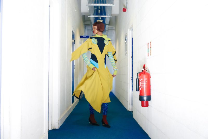 A woman in a vibrant yellow dress walks in a hallway with blue carpet, flanked by white walls and a red fire extinguisher.