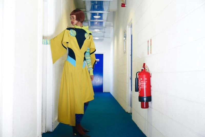 Woman in a yellow geometric dress stands in a hallway with a red fire extinguisher on the wall, creating a striking fashion editorial.