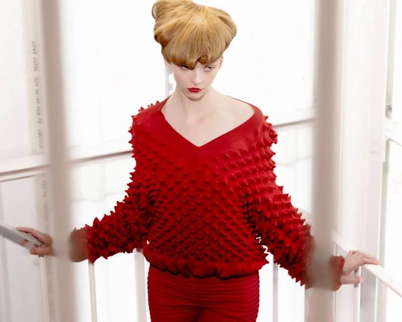 Woman in textured red top and pants poses near white railing in a high-fashion editorial shot with unique hair styling and bright lighting.