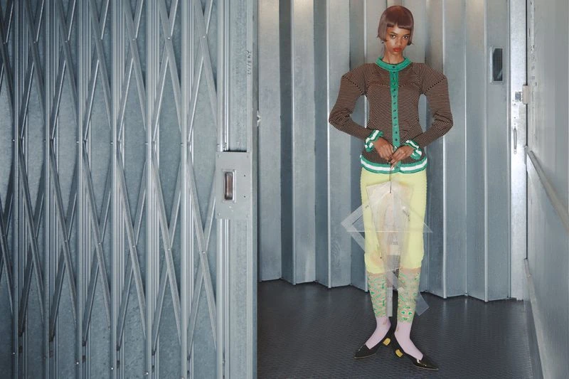 Fashion shot of an adult woman in a striped top and yellow skirt, posing indoors against a metal wall and a metal diamond-patterned gate.
