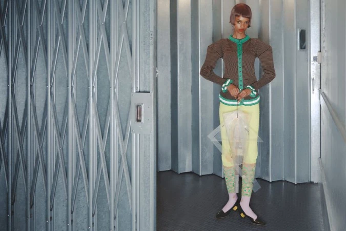 Fashion shot of an adult woman in a striped top and yellow skirt, posing indoors against a metal wall and a metal diamond-patterned gate.