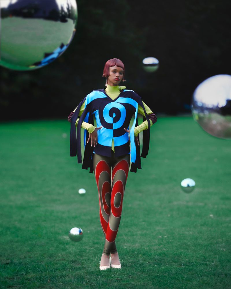 Fashionable woman poses in nature wearing unique blue, black, and yellow top with leggings; metal spheres in the grass.