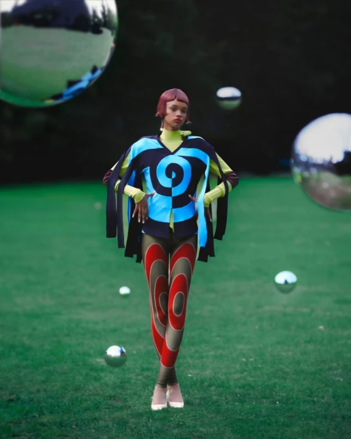 Fashionable woman poses in nature wearing unique blue, black, and yellow top with leggings; metal spheres in the grass.