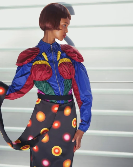 Woman with short hair posing in a colorful jacket and patterned skirt in front of a white and grey stairwell.