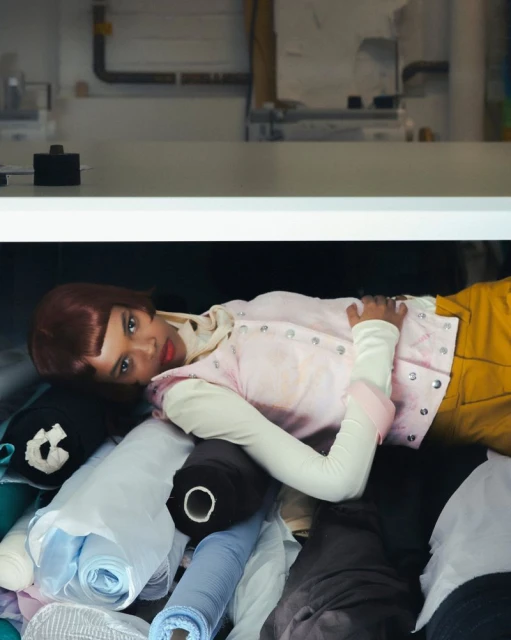 A person with a short, auburn bob hairstyle lies among rolls of fabric. They wear yellow pants and a pink vest, inside with white walls.