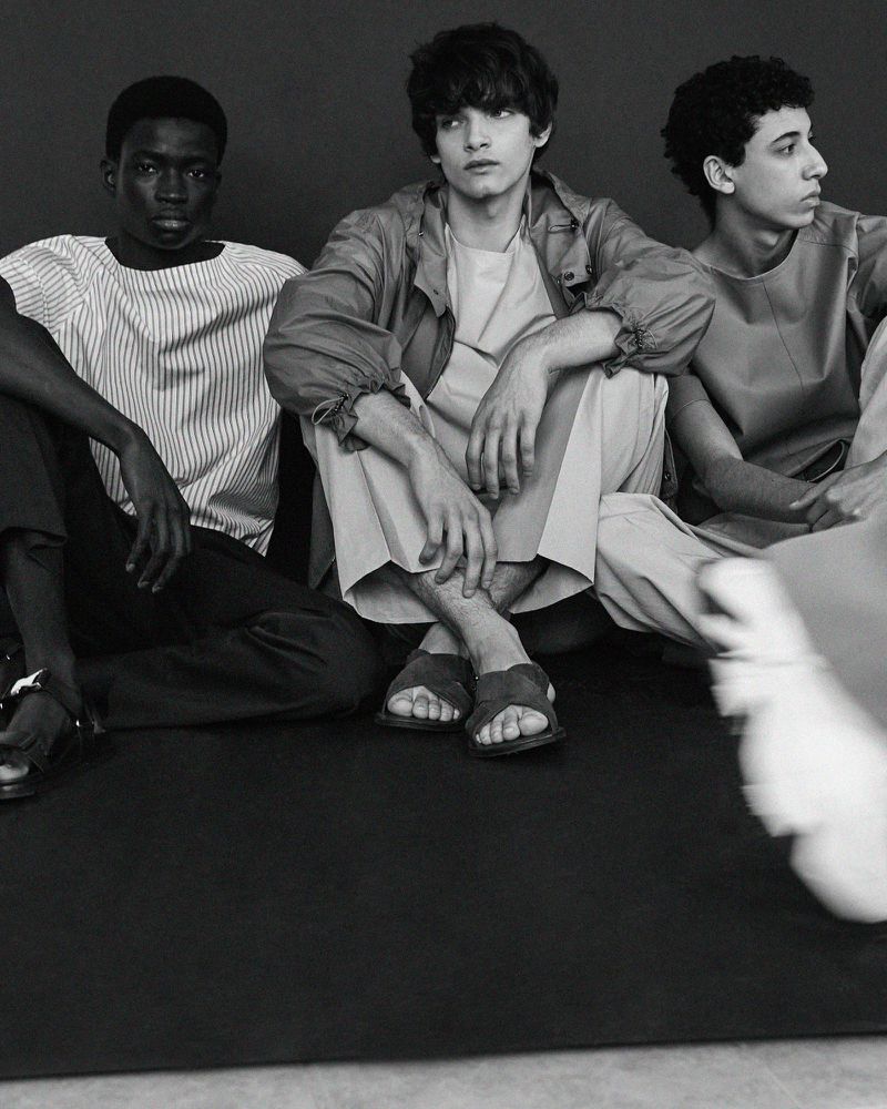 Monochrome studio shot featuring three adult male fashion models sitting on the floor wearing casual clothing, sandals, and minimalist tops.