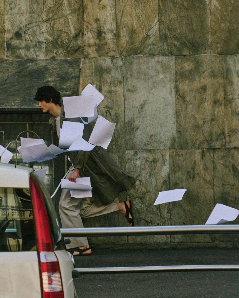 Adult male running outdoors in sandals and billowing olive clothing, scattering papers, beside a white car and metal railing.