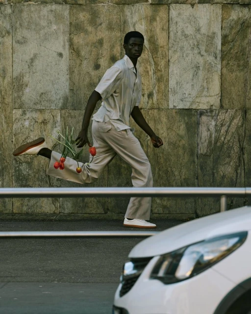 An adult male of African descent in a light-colored suit is captured in mid-stride on a city street near a car, holding tulips.