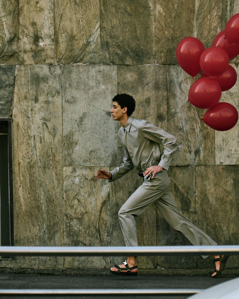 Young adult runs along city street with a bunch of red balloons. He wears a light gray suit with sandals, beside a stone wall.