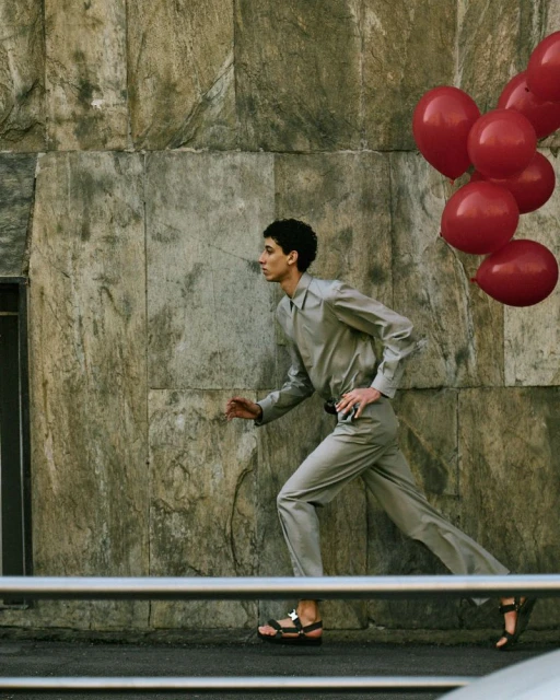 Young adult runs along city street with a bunch of red balloons. He wears a light gray suit with sandals, beside a stone wall.