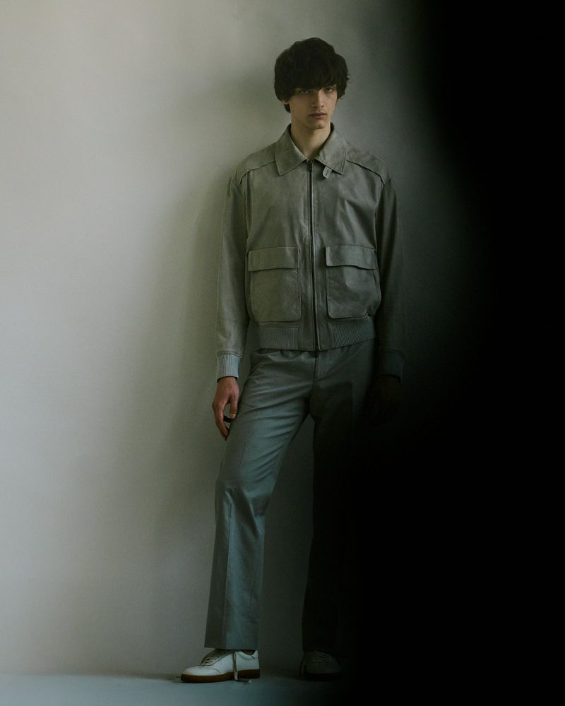 Male model poses in a grey bomber jacket and trousers, standing against a light grey wall with a partially shadowed side view.