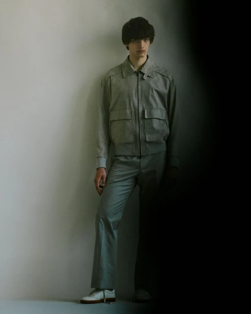 Male model poses in a grey bomber jacket and trousers, standing against a light grey wall with a partially shadowed side view.