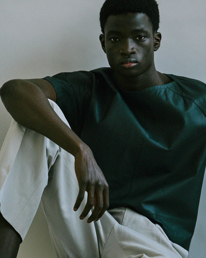 Portrait of a Black man seated wearing a dark green top and light trousers against a neutral wall.