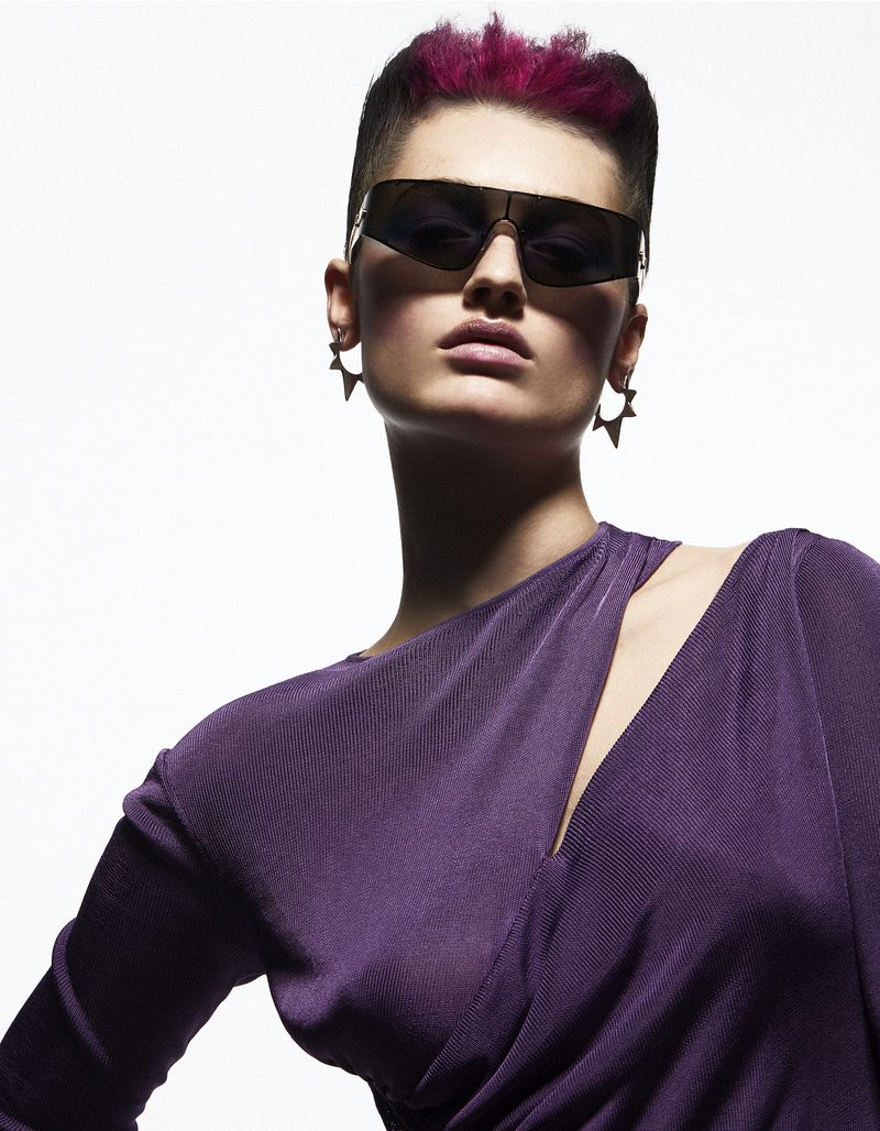 Close-up portrait of a stylish adult woman wearing sunglasses and a purple dress, posing confidently against a bright white backdrop.