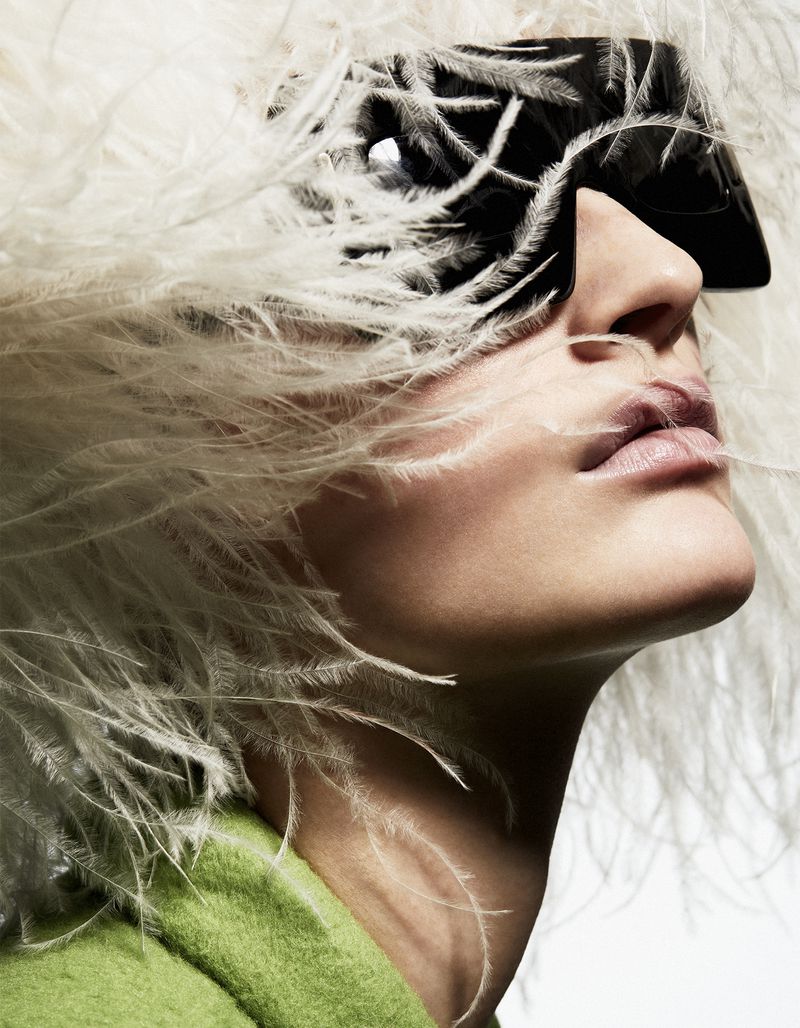 Model wearing large black sunglasses and a white feathered hat against a plain background in a fashion portrait for The Collector Magazine.