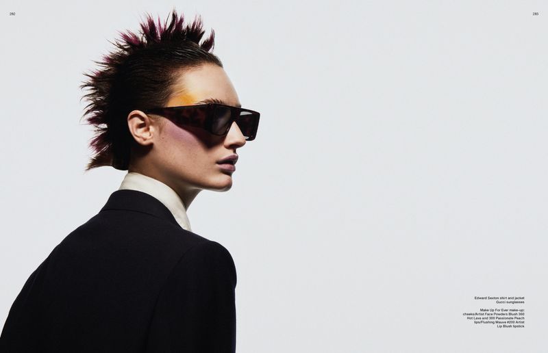 A stylish adult female in a black suit and sunglasses, with a colorful, spiky hairstyle, poses in a studio with a white background.