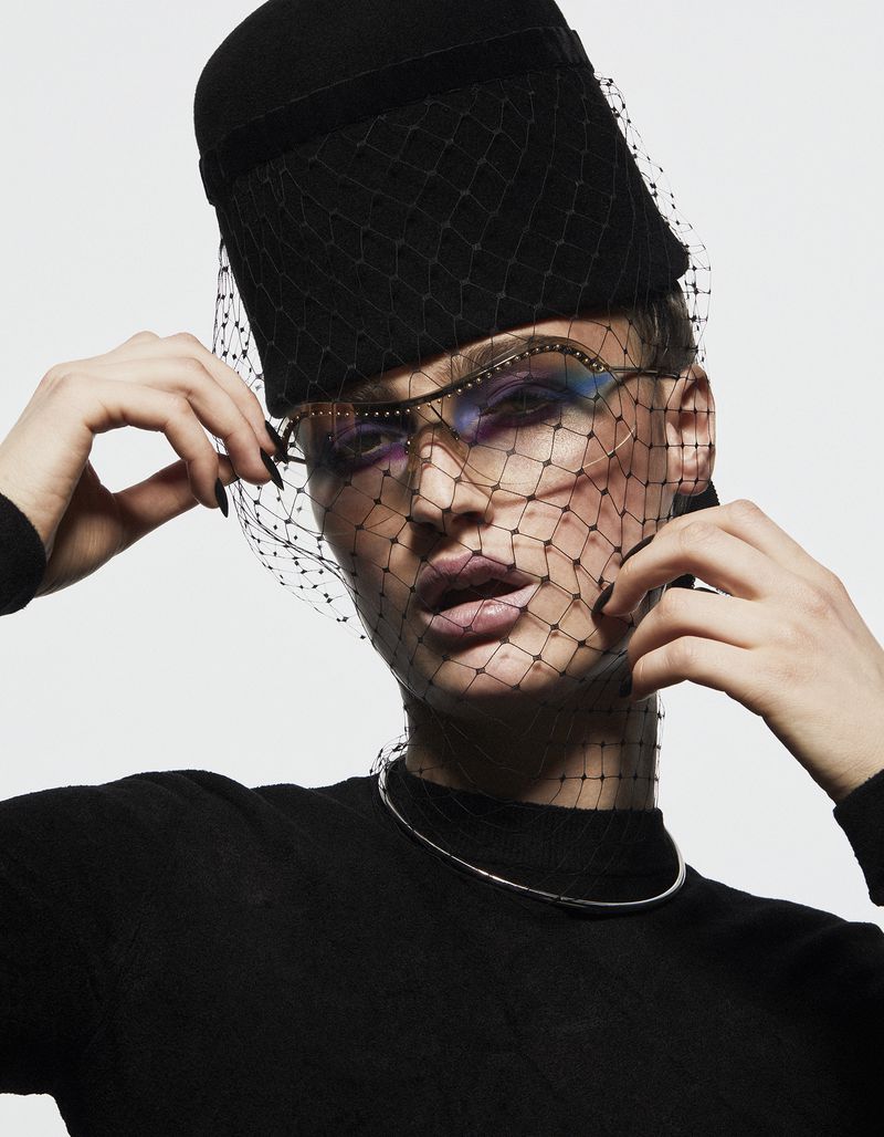 Close-up fashion portrait of a model wearing a black hat with netting, decorative glasses, and silver choker against a white background.
