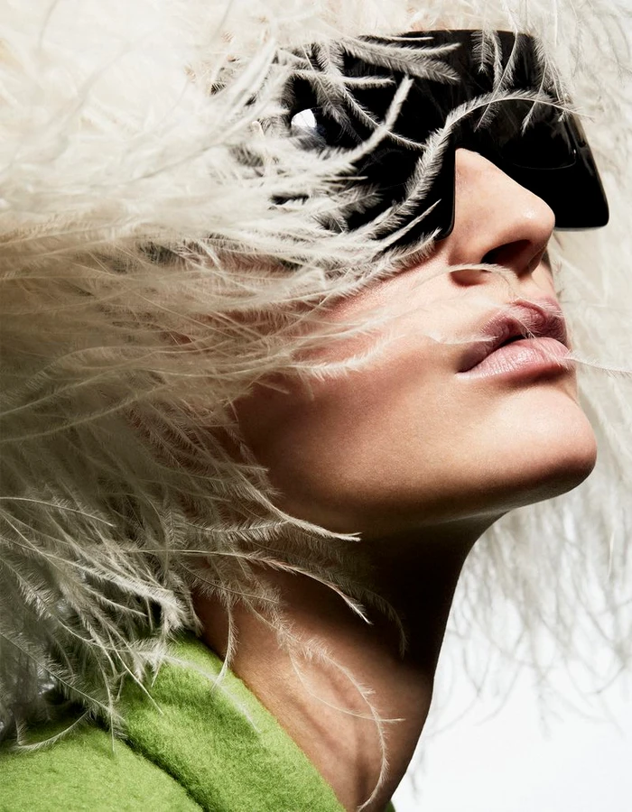 Model wearing large black sunglasses and a white feathered hat against a plain background in a fashion portrait for The Collector Magazine.