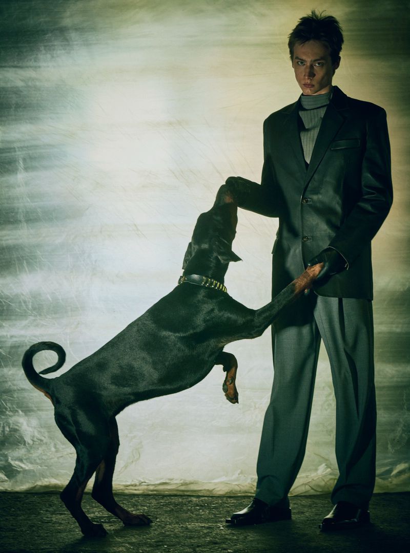 Stylish man in a suit holds a Doberman dog standing on its hind legs in a studio setting with textured background lighting.