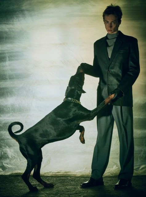 Stylish man in a suit holds a Doberman dog standing on its hind legs in a studio setting with textured background lighting.