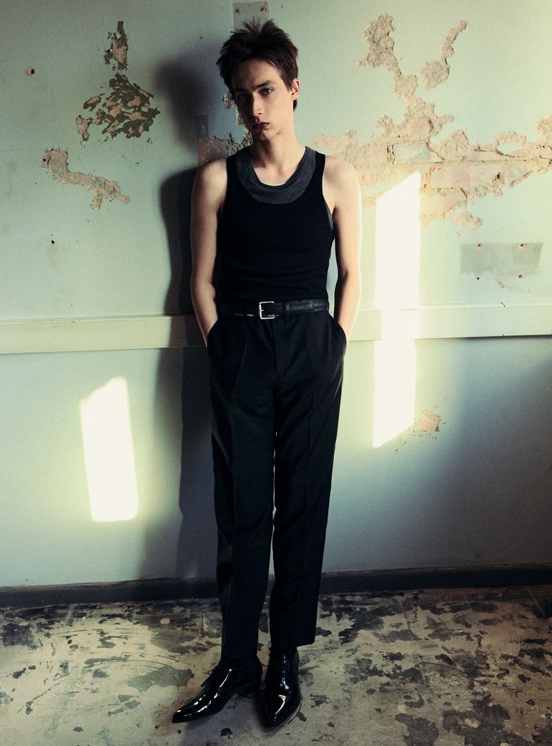 Full shot of a male model leaning against a worn wall, wearing a black tank top, black pants with a belt, and shiny black boots.