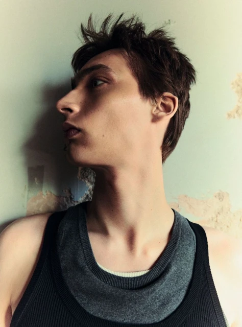 Profile of a young adult male with short brown hair wearing a layered tank top, standing indoors against a textured wall.