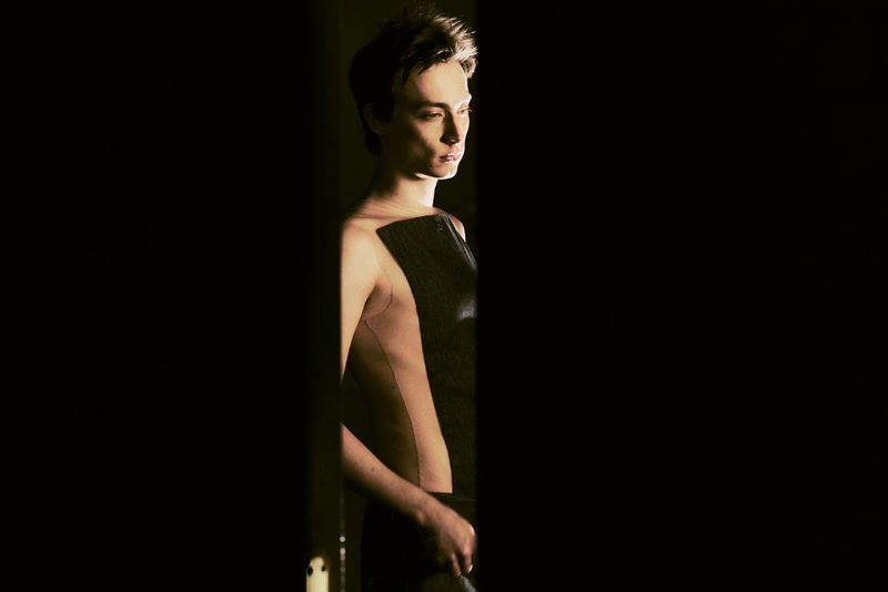 Side-lit portrait of a young adult in a partial sleeveless top, set against a black background creating a dramatic and moody atmosphere.