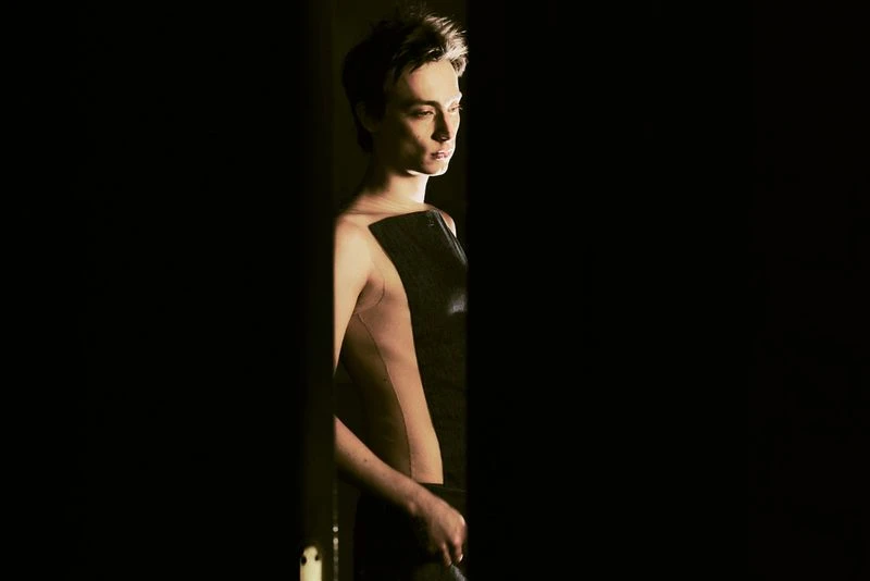 Side-lit portrait of a young adult in a partial sleeveless top, set against a black background creating a dramatic and moody atmosphere.