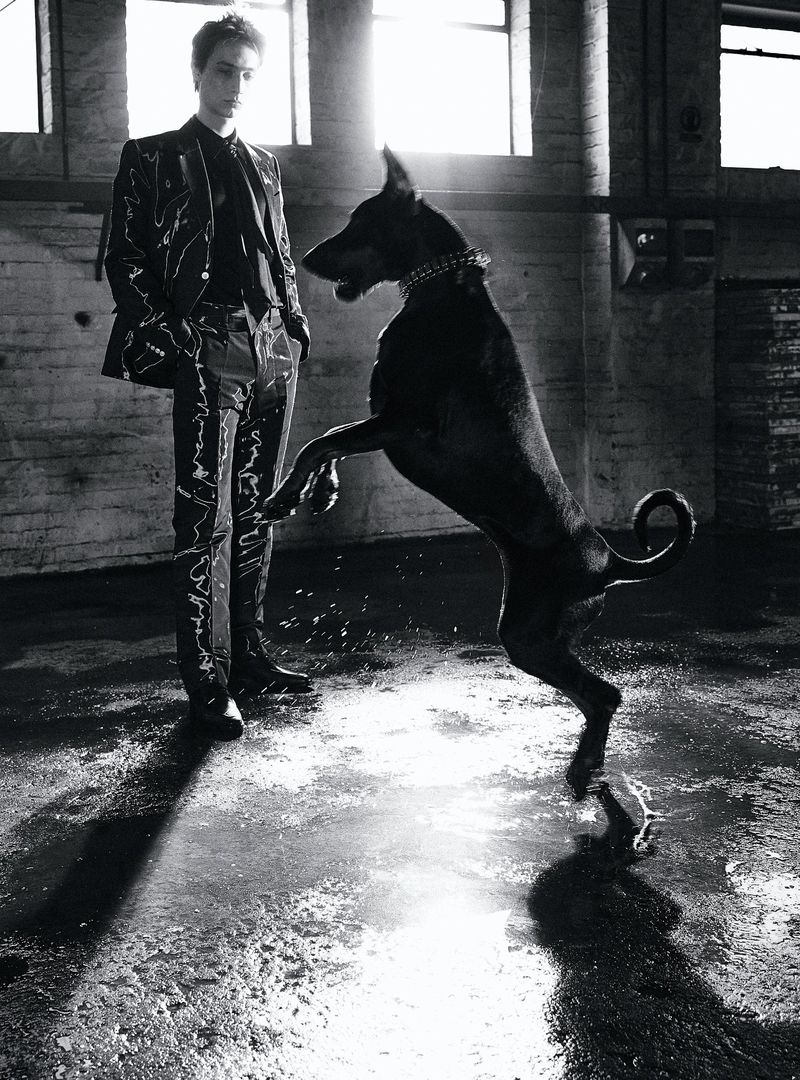 A tall, slender man in a patterned suit stands beside a large black dog on its hind legs in a high-ceilinged industrial space.