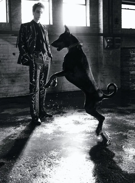 A tall, slender man in a patterned suit stands beside a large black dog on its hind legs in a high-ceilinged industrial space.