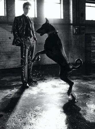 A tall, slender man in a patterned suit stands beside a large black dog on its hind legs in a high-ceilinged industrial space.