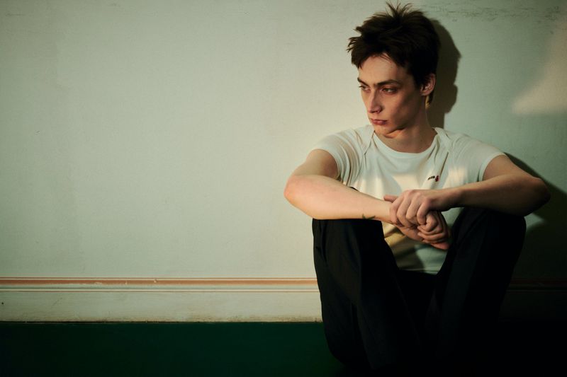 Young model in a white t-shirt and dark trousers seated on the floor against an off-white wall for The Greatest Magazine.