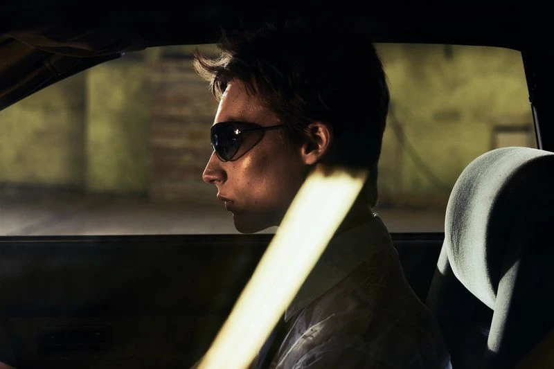 Man in sunglasses sits in car at night, illuminated by golden light. He's wearing a button-down shirt with dark interior and building backdrop.