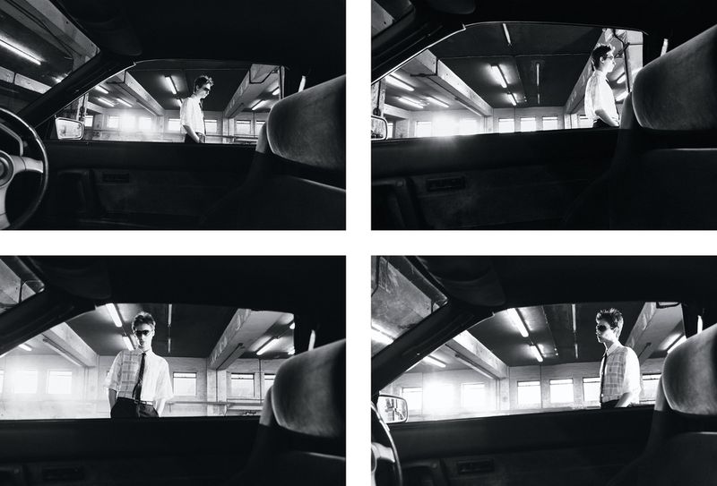Monochrome medium shot from inside a car looking out at an adult male in sunglasses standing in an urban parking garage setting.