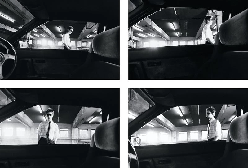 Monochrome medium shot from inside a car looking out at an adult male in sunglasses standing in an urban parking garage setting.