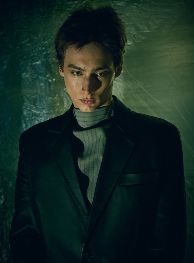 Close-up portrait of a young adult male with dark hair wearing a gray turtleneck and black blazer, with a textured green background.