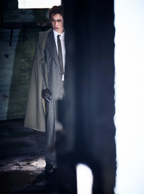 Fashionable adult male in suit with overcoat poses in dim, gritty indoor environment with dark colors. Editorial fashion shot.